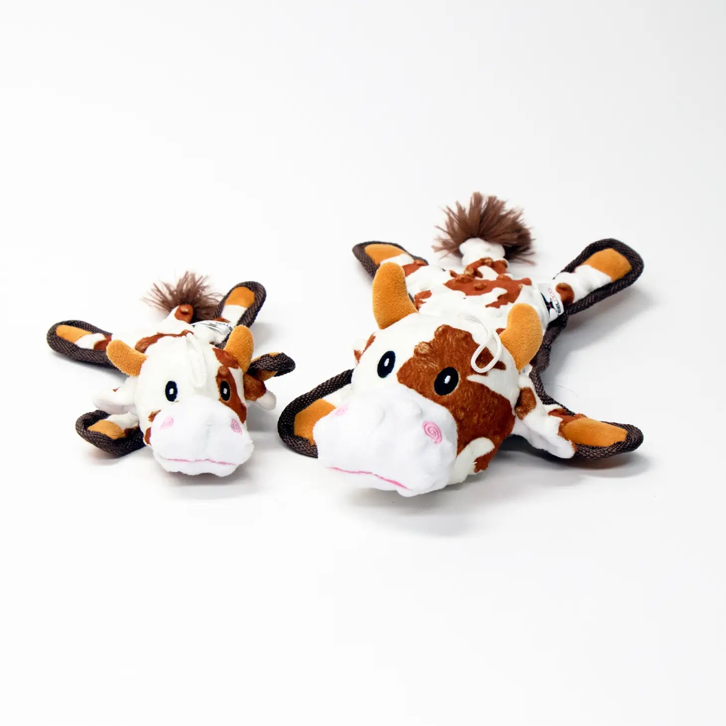 Two Bumpie Brown Cow Dog Toys laying on their stomachs. One Bumpie Brown Cow on the left is the small version of the dog toy. The Bumpie Brown Cow on the right is the standard version of the dog toy.