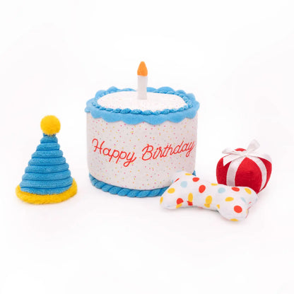 Isolated picture of the Happy Birthday cake burrow toy showcasing a small red plush present with white ribbon, a plush spiral birthday party hat and a plush white bone with multicolor polkadots